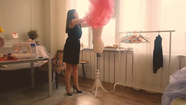 Vrouwelijke ontwerper werkt aan een roze transparante jurk in haar heldere studio. Jonge vrouw kleedt een jurk op een witte etalagepop. — Stockvideo
