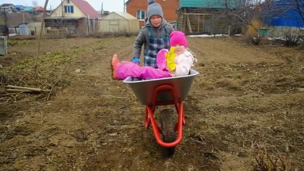 10 years old boy rides a 6 year old girl on a wheelbarrow in the country. Brother and sister are fooling around in the garden. Garden work. — Stock Video