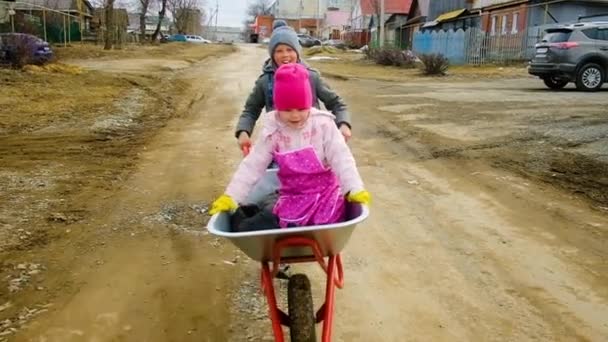 10 years old boy rides a 6 year old girl on a wheelbarrow in the country. Brother and sister are fooling around in the garden. Garden work. Riding a cart along the village street — Stock Video