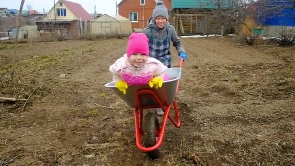 Un niño de 10 años monta a una niña de 6 años en una carretilla en el país. Hermano y hermana están jugando en el jardín. Trabajos de jardín . — Vídeo de stock