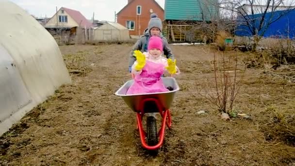 10 yaşında bir çocuk, 6 yaşında bir kızı el arabasıyla taşraya götürüyor. Erkek ve kız kardeş bahçede oynaşıyorlar. Bahçe işleri. — Stok video