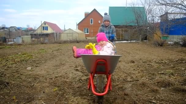 Ein 10-jähriger Junge reitet ein 6-jähriges Mädchen auf einer Schubkarre auf dem Land. Bruder und Schwester tummeln sich im Garten. Gartenarbeit. — Stockvideo