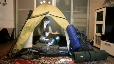 Dairesinde kamp çadırında maskeli bir kız kitap okuyor. Evde kendini izole etme. Koronavirüs salgını. Seyahat etmeyi ve gezmeyi özlüyor.