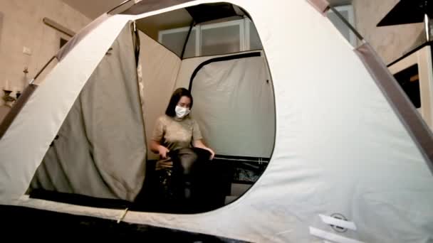 Mädchen in medizinischer Maske legt in einem Zelt in einer Wohnung einen Schlafsack aus. Selbstisolierung zu Hause. Coronavirus-Pandemie. Reisen und Wandern verpasst — Stockvideo