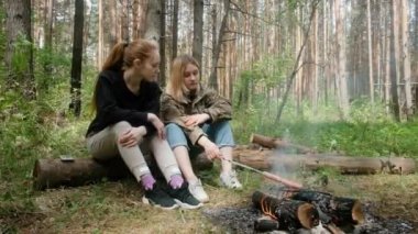 İki genç kız ormanda ateşin etrafında sosis kızartıyorlar..
