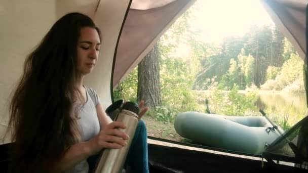 Young woman tourist is sitting in a tent and drinking drinks from a thermos. — Stock Video