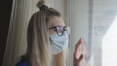 Tıbbi koruyucu maskeli bir kadın pencereden dışarı bakar..