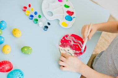 Çocuk, beyaz bir odada masa başında oturan ahşap yumurta parçalarına yumurta boyuyor.