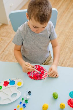 Çocuk, beyaz bir odada masa başında oturan ahşap yumurta parçalarına yumurta boyuyor.