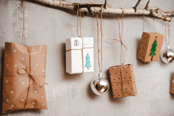 Regalos de Navidad Embalado con papel Kraft y decoraciones hechas a mano cuelgan de una cuerda atada a un palo contra una pared de hormigón gris. — Foto de Stock