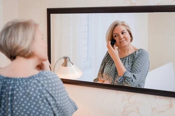 Feliz de mediana edad anciana mujer preens y ajusta su cabello delante del espejo del tocador — Foto de Stock