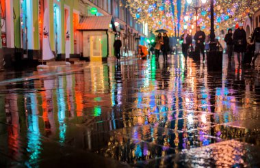 Büyük bir şehirde yağmurlu bir gece, ıslak yol yüzeyindeki renkli şehir ışıklarının yansıması. Aydınlık şehir tatili aydınlatmalı bir yaya sokağı manzarası.