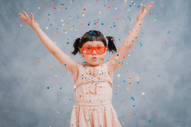 Küçük kız neşeli bir şekilde gri mavi arka plana renkli süsler ve konfeti kusar. Tatil günü. Mutlu, heyecanlı, gülen bebek doğum gününde.