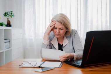 Yaşlı yaşlı kadın, iş kararından dolayı kafası karışmış kağıt belgelerle bilgisayarda boğuşuyor..