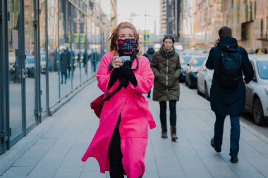 Parlak pembe paltolu kadın, yüzünde virüsün bulaştığı havadan etkilenen şık, modaya uygun koruyucu bir maske takmış kameralı bir turist. Rusya Moskova 10 Mart 2020.