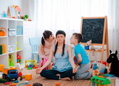 Anne zemin katta oturan çocuklar için yemek odası oyuncakları, meditasyon pozisyonu, küçük çocuklar çığlık çığlığa etrafta oynuyorlar odaklanmış anne meditasyon yapıyor, oyuncu çocuğu görmezden geliyor. Stressiz kavramlar