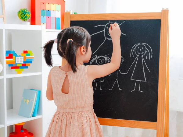 Uma menina fica no quarto das crianças com um quadro-negro e desenha uma família com giz . — Fotografia de Stock