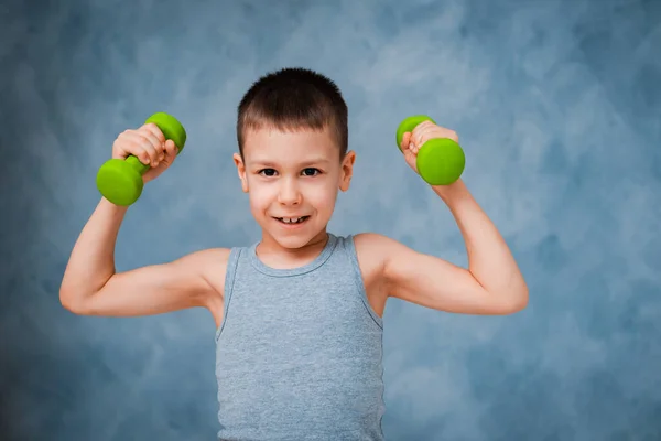 Cry sports baby boy emotionally shows his arm bicep muscle strength with dumbbells grey blue background — Stock Photo, Image