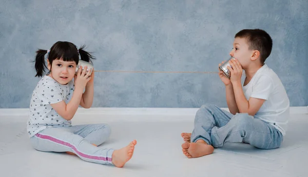 A young brother and sister are talking to a tin phone against a gray wall. — Stock Photo, Image