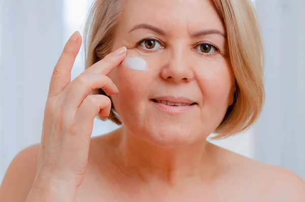 senior woman anti-aging lotion against dark circles under the eyes. Happy Mature woman uses cosmetic cream to hide wrinkles. A lady using a day moisturizer.
