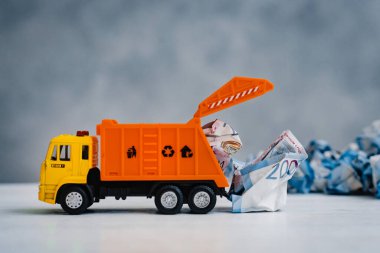 Turuncu oyuncak çöp kamyonu mavi arka plandaki beyaz bir masaya buruşmuş kağıt banknotları boşaltıyor. Para çöplüğü, boş alan