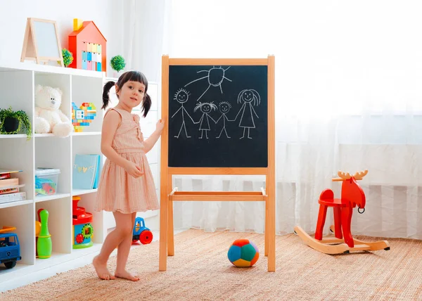 一个女孩拿着黑板站在孩子们的房间里 用粉笔画了一个家庭 家庭和幼儿园游戏的概念 — 图库照片