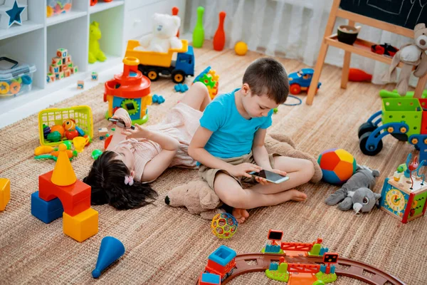 Hermanos niños hermano y hermana, amigos se sientan en el suelo de la casa en la sala de juegos para niños con teléfonos inteligentes, separados de los juguetes dispersos . —  Fotos de Stock
