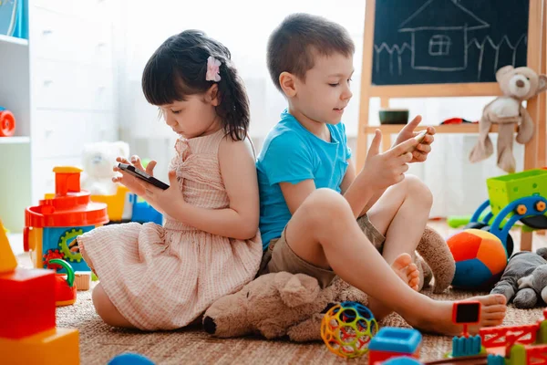 Frères et sœurs enfants frère et sœur, amis assis sur le sol de la maison dans la salle de jeux pour enfants avec smartphones, détachés des jouets éparpillés . — Photo