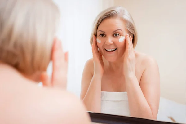 Retrato mujer bastante mayor con las manos en el espejo de la cara en casa después del baño cuidado de la piel después de 50-60 años — Foto de Stock