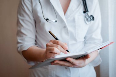 Kadının el doktoru reçeteli bir konsültasyon formu hazırlıyor. Beyaz önlüklü ve camında steteskop olan bir tıp çalışanı bir günlüğe notlar alır.