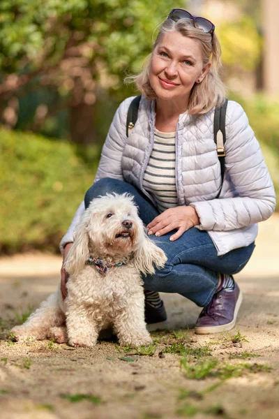 Yetişkin sarışın kadın şehir parkında tüylü beyaz köpekle yürüyor. — Stok fotoğraf