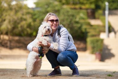 Yaşlı emekli kadın parkta küçük beyaz köpek gezdiriyor.
