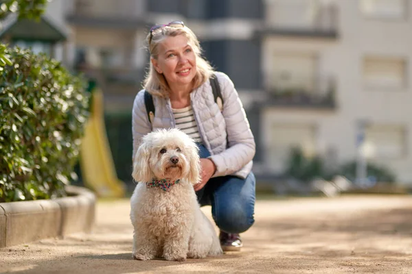 Medelålders blond kvinna promenader med fluffig vit hund i sommar stad — Stockfoto