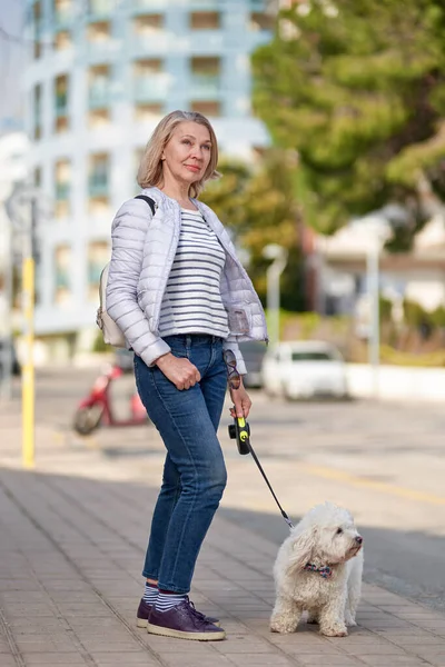 Attraktiv medelålders blond kvinna promenader med fluffig vit hund i sommar stad — Stockfoto