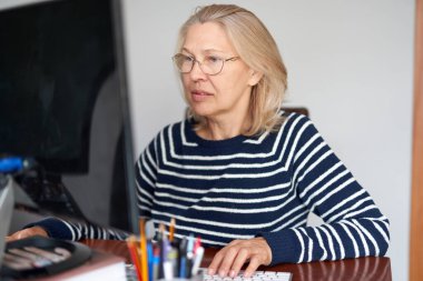 Yetişkin orta yaşlı ve gözlüklü. Ev ofisinde çalışan dizüstü bilgisayarlı e-posta kullanıyor. İnternette bilgi arayan ya da internetten iletişim kuran kadın.