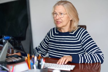 Yetişkin orta yaşlı ve gözlüklü. Ev ofisinde çalışan dizüstü bilgisayarlı e-posta kullanıyor. İnternette bilgi arayan ya da internetten iletişim kuran kadın.