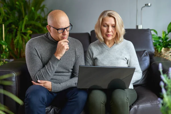 Elderly mother sit rest on sofa with grown-up son watch video on modern laptop,middle-aged mom rest in living room with adult child use computer family weekend together