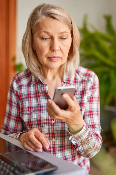 Volwassen vrouw met een telefoon in haar hand op zoek naar laptop — Stockfoto