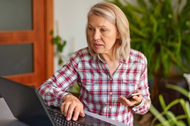 Elinde telefonla dizüstü bilgisayara bakan olgun bir kadın.