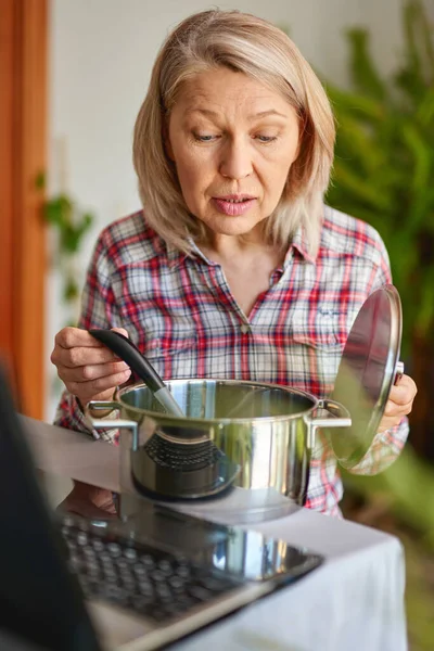 Cooking woman looking at computer while preparing food in kitchen. Beautiful mature woman reading cooking recipe