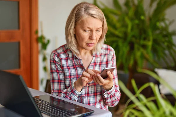 Mulher madura com um telefone na mão olhando para laptop — Fotografia de Stock