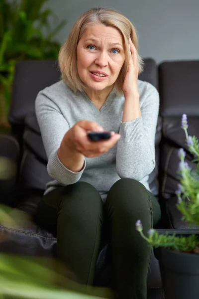 Volwassen vrouw thuis zitten op de bank en tv kijken, ze houdt een afstandsbediening. — Stockfoto