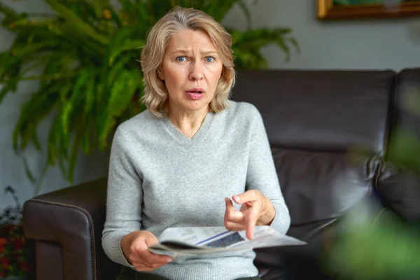 Porträt einer besorgten Frau, die schlechte Nachrichten in der Zeitung liest und zu Hause auf einem Sofa im Wohnzimmer sitzt. — Stockfoto