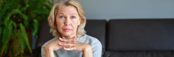 Blondynka w średnim wieku siedzi na kanapie w domu. — Zdjęcie stockowe