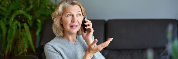 Gelukkig volwassen vrouw met behulp van mobiele telefoon terwijl zitten op de bank thuis. — Stockfoto