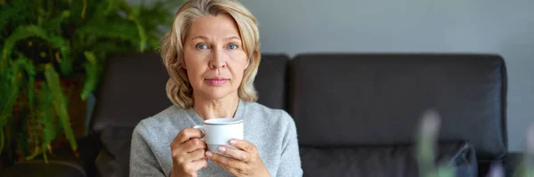Heerlijk drank.Oudere vrouw zit op de bank in haar woonkamer en houdt een kopje koffie vast. — Stockfoto