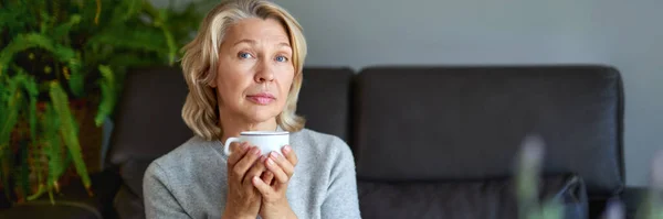 Lahodné pití. Starší žena sedí na pohovce ve svém obývacím pokoji a drží šálek kávy. — Stock fotografie