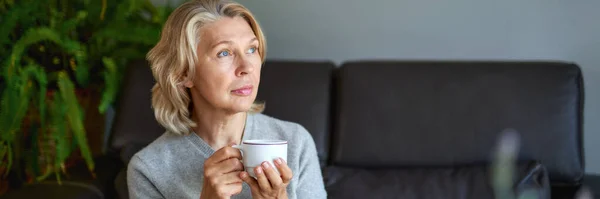 Bebida deliciosa.Mulher idosa sentada no sofá em sua sala de estar e segurando uma xícara de café . — Fotografia de Stock