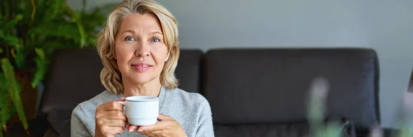 Mulher idosa alegre sentado no sofá em sua sala de estar e segurando uma xícara de café — Fotografia de Stock