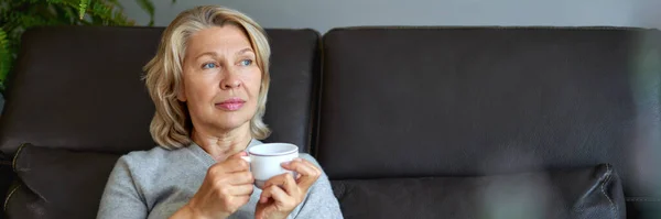 Mulher idosa alegre sentado no sofá em sua sala de estar e segurando uma xícara de café — Fotografia de Stock